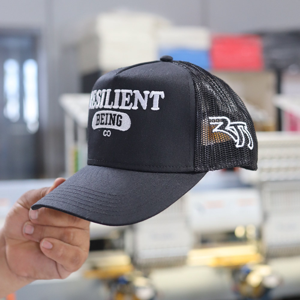 A man holding the front view of a OTTO CAP 32-285 5 Panel Mid Profile Mesh Back Trucker Hat featuring the custom embroidery of "Resilient Being Co." logo in bold design embroidered in white thread by Garment Decor