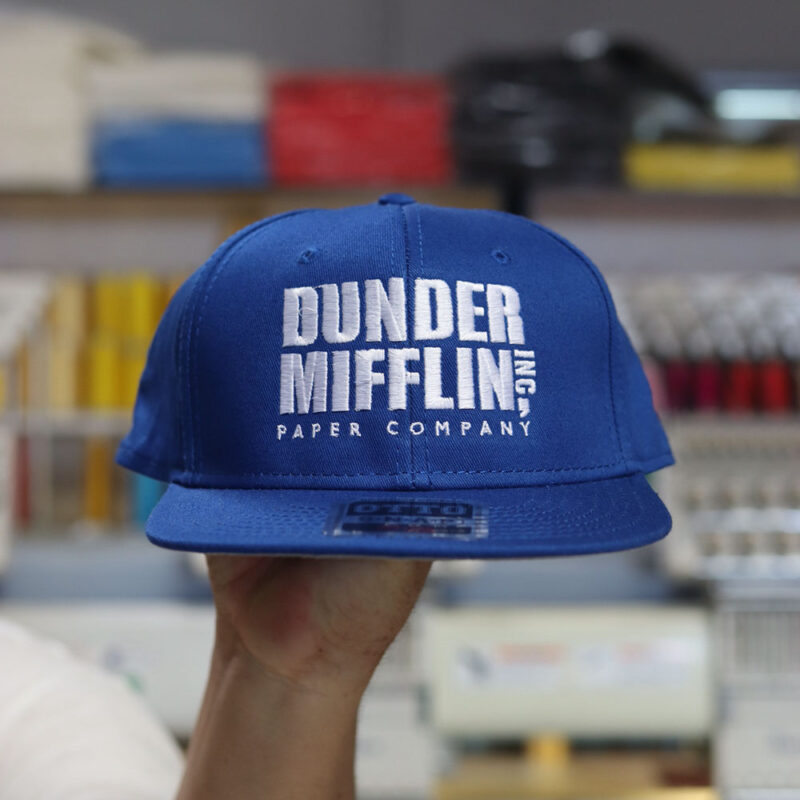 A man holds the front of a royal blue OTTO CAP 125-1038 "OTTO SNAP" 6-panel mid-Profile Snapback Hat, which showcases the custom embroidery of the "Dunder Mifflin Inc. Paper Company" logo. Expertly embroidered by Garment Decor, the embroidery highlights precise detailing and vibrant white threadwork for a polished finish.