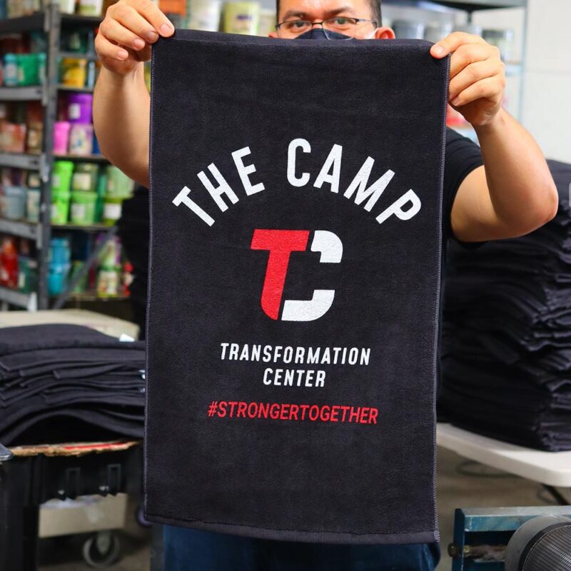 This image features Garment Decor's custom screen printed black towel designed for "The Camp Transformation Center". The towel displays a bold, white arched text "The Camp," with a striking red and white "T C" logo beneath it. The phrase "Transformation Center" is printed in white, followed by the hashtag "#StrongerTogether" in red. The towel is held by a person in a workshop filled with vibrant printing supplies and folded towels in the background. Perfectly designed for promotional or fitness branding purposes.