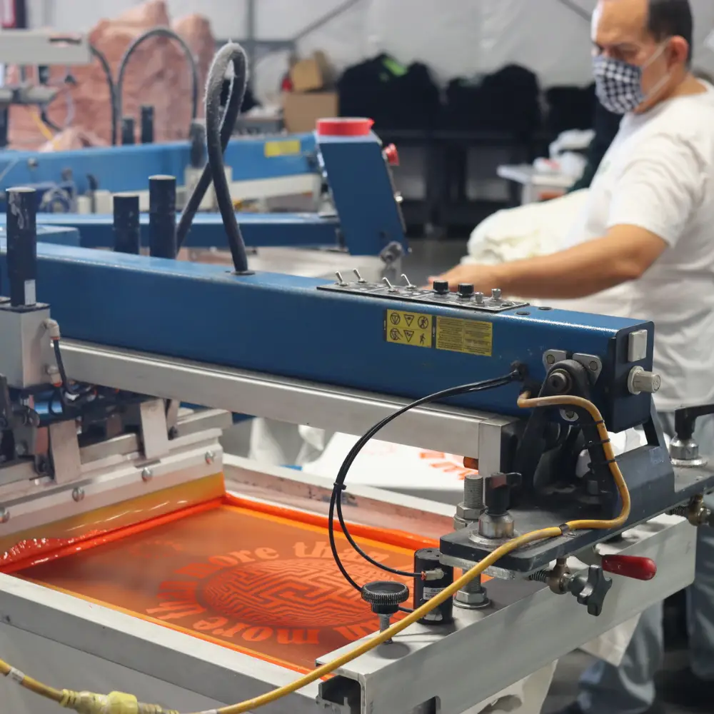 The Garment Decor screen printer expert carefully setting up the Los Angeles Apparel 1801GD Garment Dye Crew Neck T-Shirt in white on an M&R machine palette for the Custom Puff Screen Print with More Than design by Uninterrupted in orange ink. This step highlights Garment Decor's precision in preparing garments for custom puff screen printing, ensuring high-quality results.
