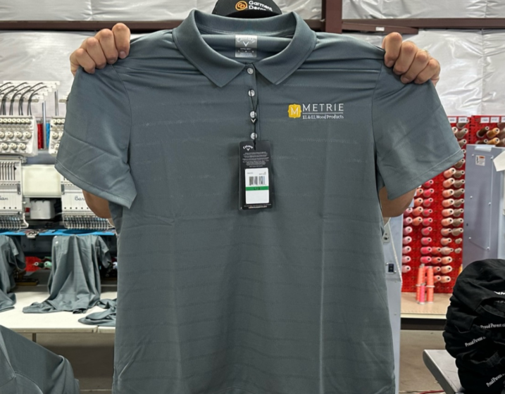 This is a photo of a gray polo that Garment Decor custom embroidered with a logo on the left chest and is being held up by our embroidery operator.