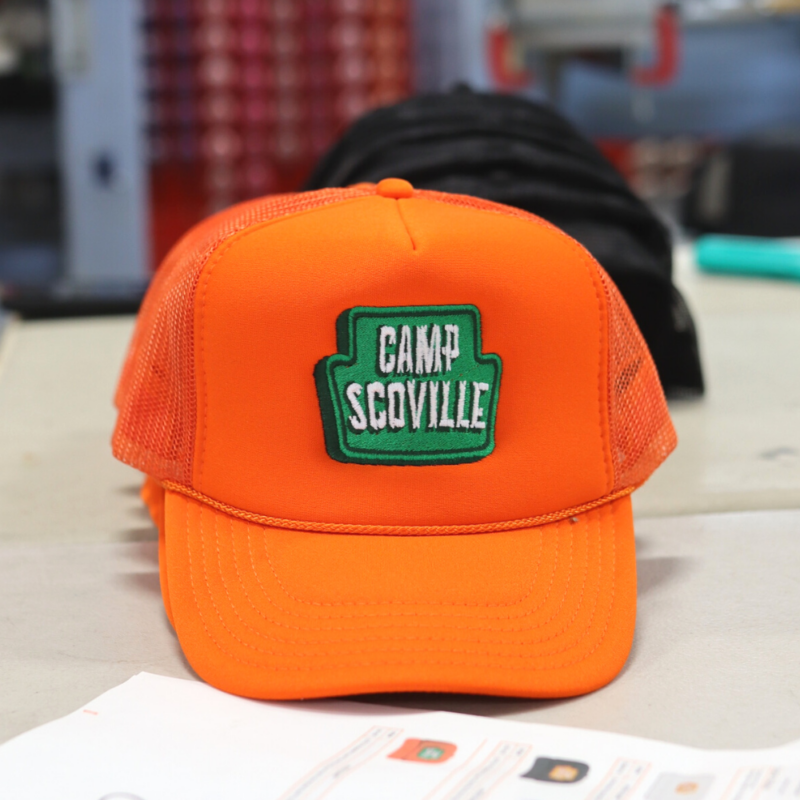 The OTTO CAP 39-165 5 Panel High Crown Mesh Back Trucker Hat in Neon Orange features a bold Camp Scoville design custom embroidered by Garment Decor. The embroidery showcases intricate stitching of a retro-inspired campfire logo in a warm green and white thread that reads "Camp Scoville". The textured mesh back complements the durable front panel, providing a stylish and breathable fit.