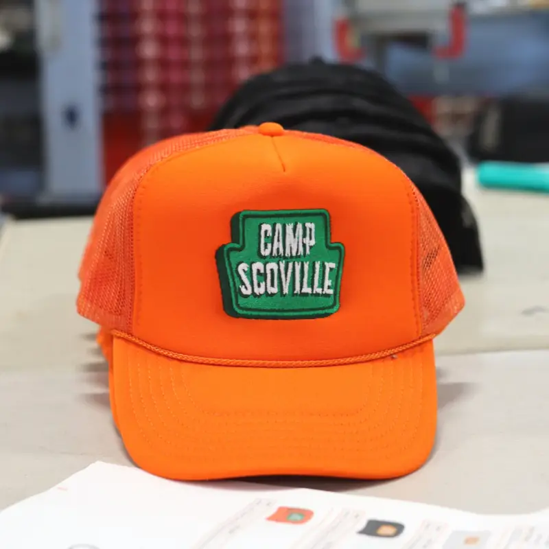 The OTTO CAP 39-165 5 Panel High Crown Mesh Back Trucker Hat in Neon Orange features a bold Camp Scoville design custom embroidered by Garment Decor. The embroidery showcases intricate stitching of a retro-inspired campfire logo in a warm green and white thread that reads "Camp Scoville". The textured mesh back complements the durable front panel, providing a stylish and breathable fit.