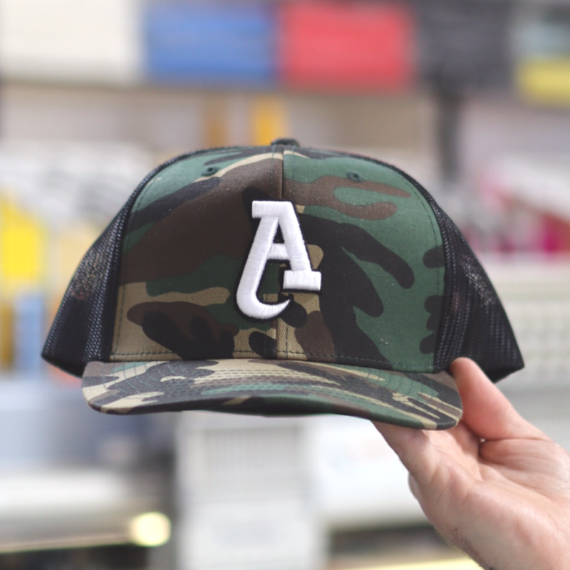 A man showcases the front view of OTTO CAP 141-1070 6 Panel Pro Style Mesh Back Trucker Snapback Hat in a striking Dark Green Camo, Brown, and Black color combination, embroidered by Garment Decor. The design features a bold capital "A" in custom 3D embroidery, with a sleek, raised finish that adds depth and texture to the hat's front panel. The earthy tones of the hat complement the intricate stitching, highlighting the precision and high-quality craftsmanship of the embroidery. The structured crown and curved bill give the hat a modern, professional look.