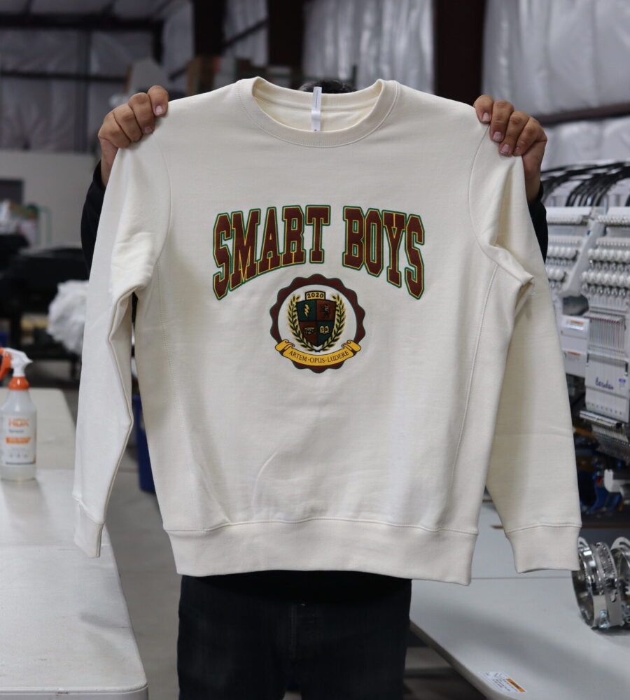 A man holds the front view of Original Favorites GOTS® Organic Cotton Crewneck Sweatshirt in natural, showcasing its dual decoration expertly crafted by Garment Decor. The front of the sweatshirt features a minimalist yet eye-catching combination of custom screen printing and embroidery. The "Smart Boys" text design is screen printed across the chest in bold maroon ink, using a clean and modern university font that contrasts elegantly against the natural-colored organic cotton fabric. Below the printed text, the Smart Boys logo is custom embroidered in vibrant maroon and yellow threads, adding texture and dimension to the design. The combination of screen printing and embroidery highlights the premium craftsmanship and unique style of this sustainably made crewneck sweatshirt.