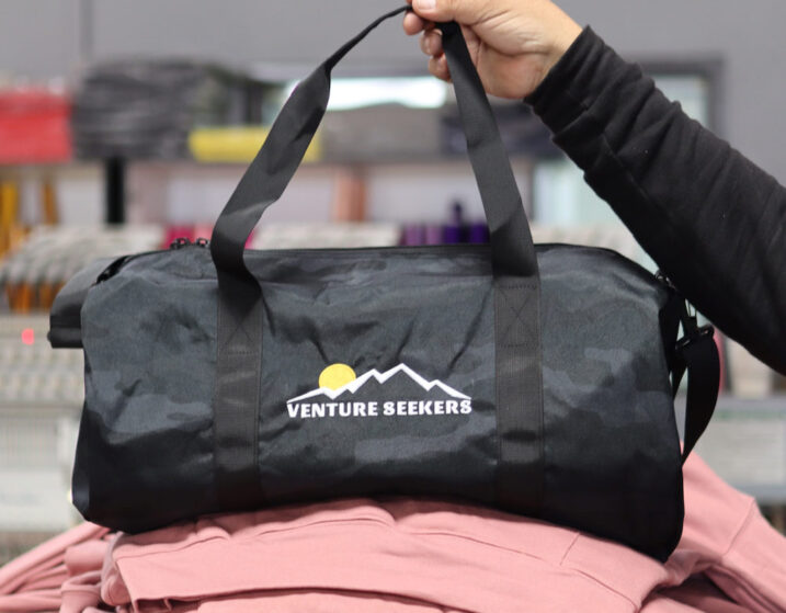 A hand holding up a black duffel bag with the logo "venture seekers" over a stack of pink fabric.