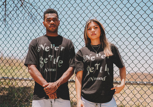 Two people standing in front of a chain-link fence wearing black t-shirts with the phrase "never settle" printed on them.