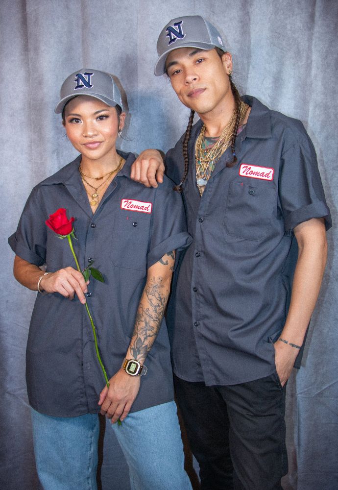 Two individuals in matching grey shirts and caps posing for a photo, one holding a red rose.