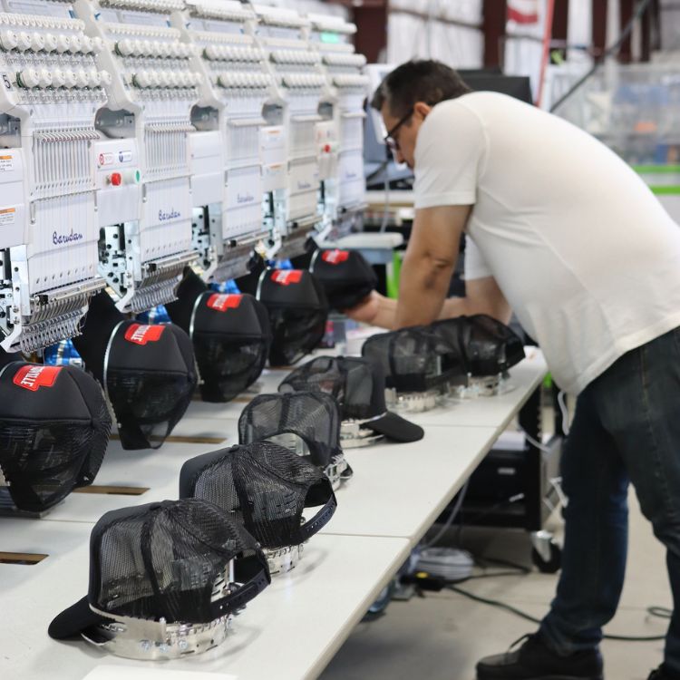Man operating embroidery machinery to customize black caps.