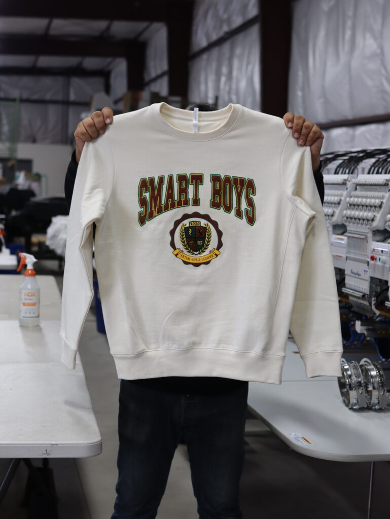 Person holding up a cream-colored sweatshirt with "smart boys" printed on it in a workshop setting.