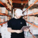 A man in a black garment-decor t-shirt and white cap using a tablet in a warehouse with rows of shelves filled with boxes.