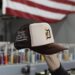 A hand holding a brown and beige trucker hat with an ornate "D" logo and text "Bridge the Gap, Build the Connection," in front of an American flag. The background is blurred with colorful spools of thread, showcasing a unique addition to our portfolio.