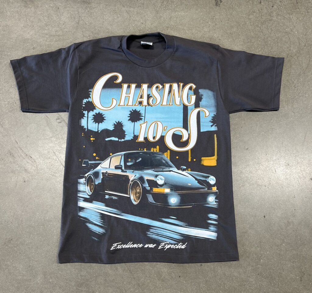 A black t-shirt with a graphic of a porsche car and palm trees, featuring the text "chasing 10's" and "excellence over epochs".