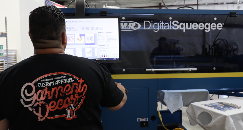 A person operates a printing machine labeled "Digital Squeegee" using a computer. The person is wearing a black shirt with "Custom Apparel Garment Decor" printed on the back.