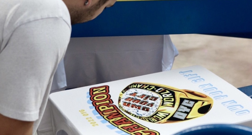 A person in a white shirt looks at a large printed image of a "World Champion" ring on a t-shirt, with the year 2022 and other text visible.