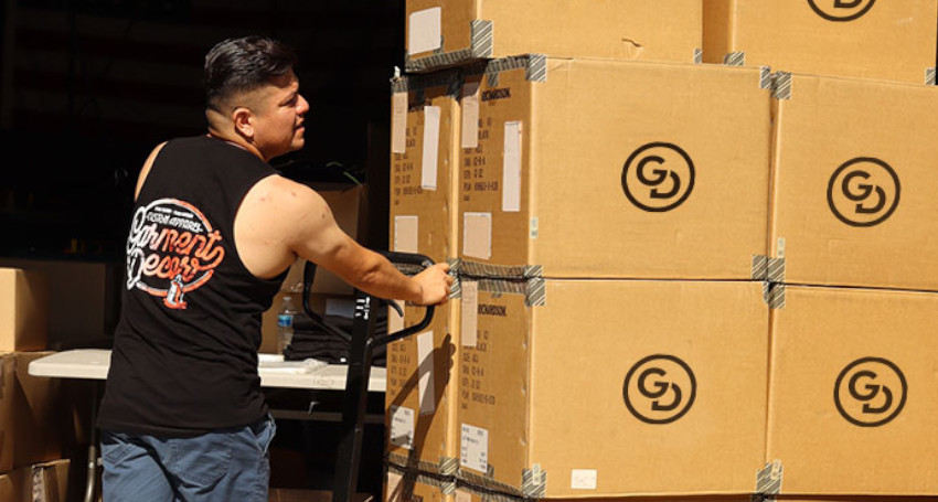 A person in a sleeveless shirt uses a pallet jack to move a stack of large cardboard boxes marked with a circular logo.