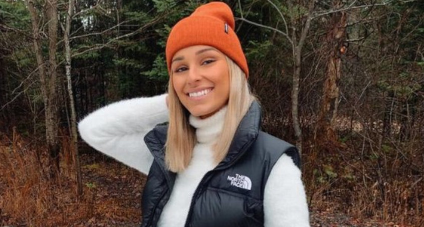 A person smiles outdoors wearing an orange beanie, white sweater, and one of the Women's The North Face Vests. Trees and foliage are visible in the background.