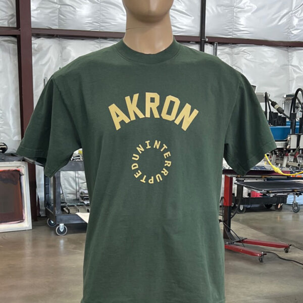 A mannequin is wearing one of the 100 Garment-Dyed Heavyweight Shirts - Text-Based Artwork with Vector Designs - Maximum of 2 Ink Colors - Soft Hand Screen Printing, featuring a green T-shirt with "AKRON" printed in large yellow letters across the front, inside a circular design. The background shows an industrial setting with machinery and workstations.