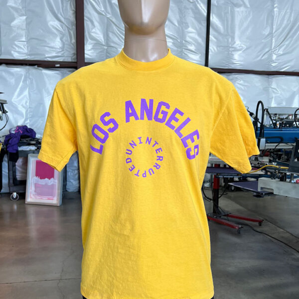 A mannequin is dressed in a 100 Garment-Dyed Heavyweight Shirt with Text-Based Artwork with Vector Designs - Maximum of 2 Ink Colors - Soft Hand Screen Printing in yellow, featuring the text "LOS ANGELES DOUNITE INT'L CREW" in purple. The mannequin is located in an industrial setting.