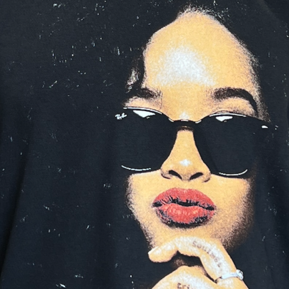 An image of a person wearing dark sunglasses and red lipstick, depicted in a print on a black T-shirt. The individual is shown resting their chin on their hand with fingers adorned in rings.