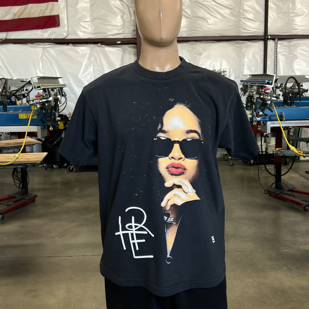 A mannequin dressed in a black T-shirt featuring an image of a person wearing sunglasses, standing in an industrial workshop with various machines in the background.