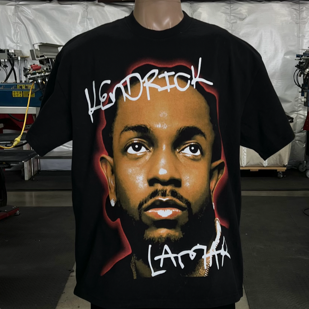 Black t-shirt featuring a large, stylized portrait of a man with Kendrick Lamar written in graffiti-style text. The background includes machinery and equipment.