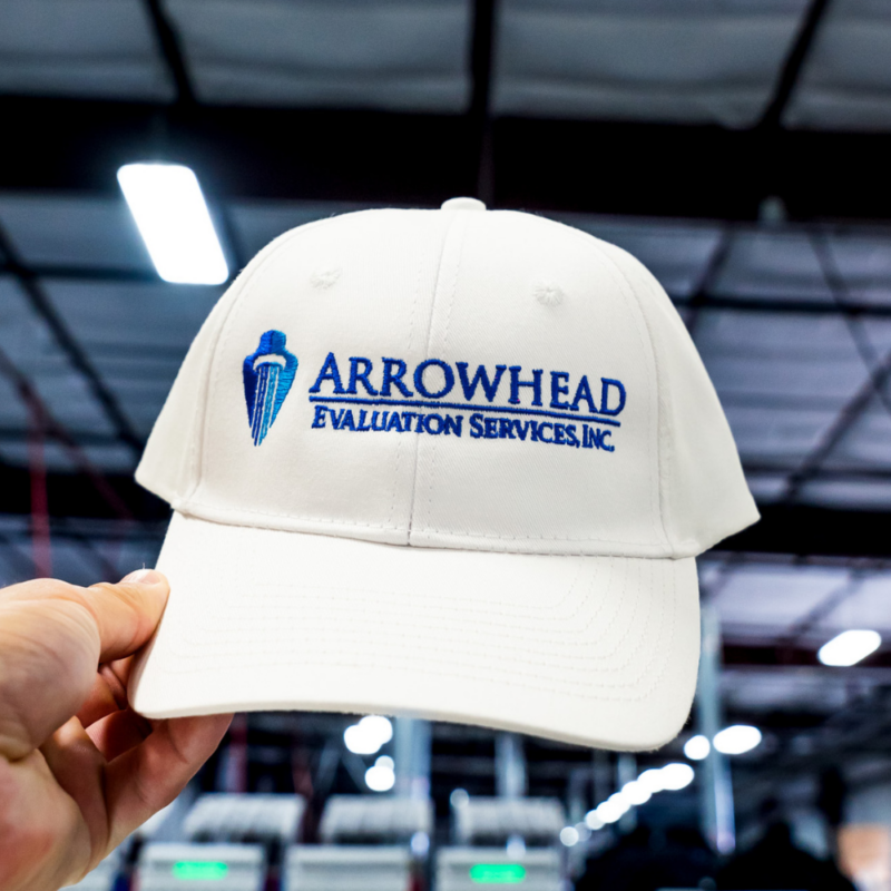 A man holding the front view of a custom embroidered Arrowhead Evaluation Services, Inc. logo on a white Valucap VC600 Chino hat by Garment Decor.