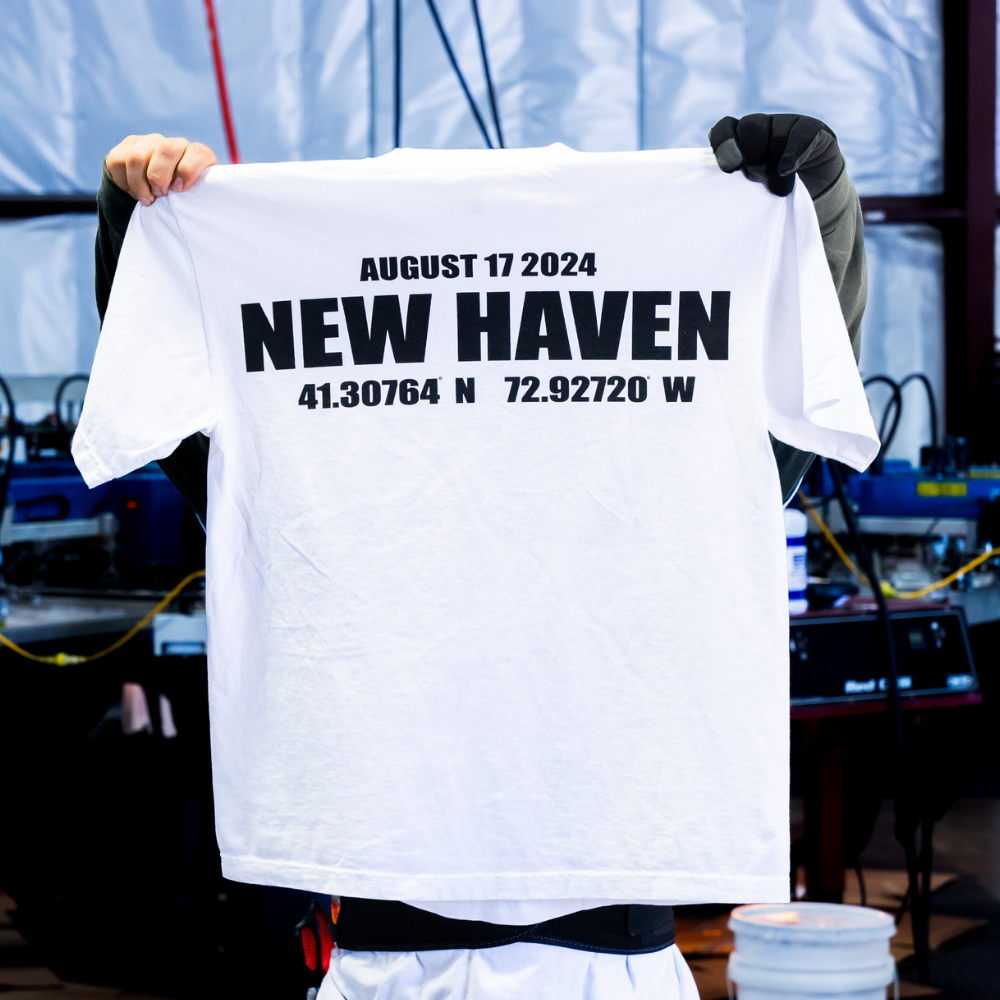 A man holding this back view highlights the smooth and professional finish of the Black Wall Street T-Shirt, printed by Garment Decor on a white Los Angeles Apparel 1801GD Garment Dye Crew Neck, ensuring style and quality for New Haven Festivals Inc.