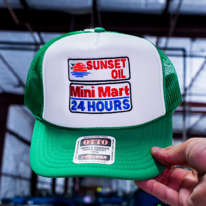 A man holding a detailed front view of the Otto Cap 39-165 5-panel high crown mesh back trucker hat in Kelly-White-Kelly, showcasing Garment Decor's custom embroidery services with a Sunset Oil Mini Mart logo.